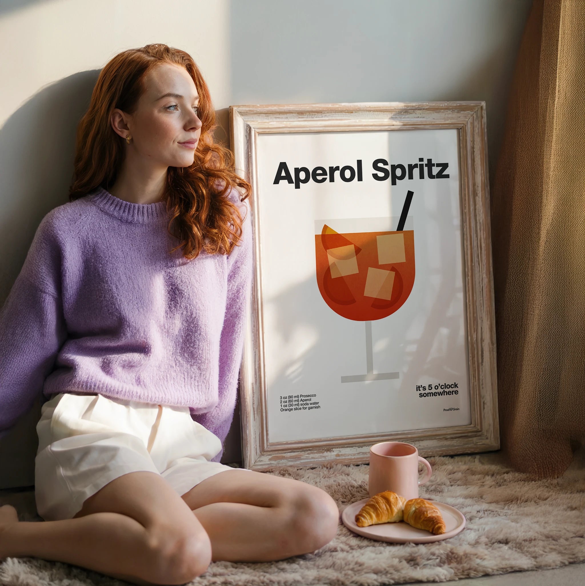 Woman sitting on a couch next to a framed Aperol Spritz poster with a plate of croissants and a mug.