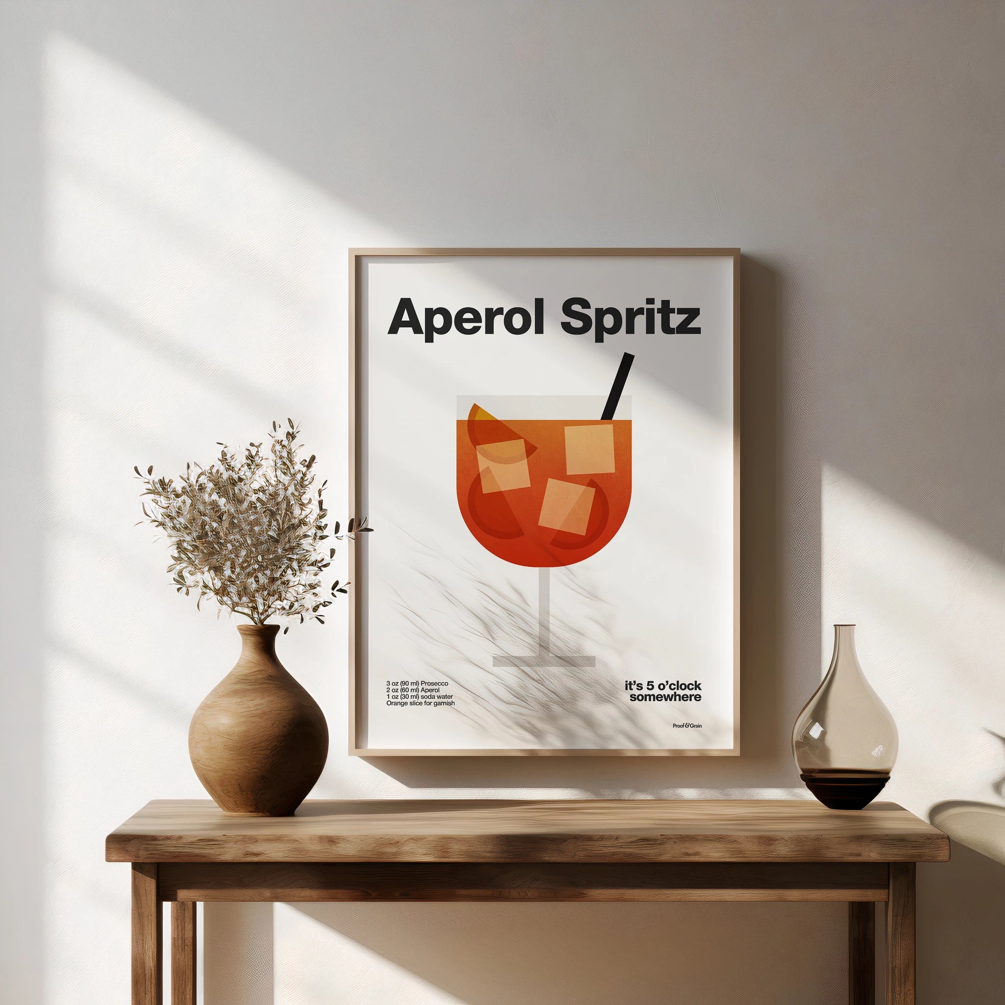 Framed Aperol Spritz poster on a wall with decorative vases on a wooden table.