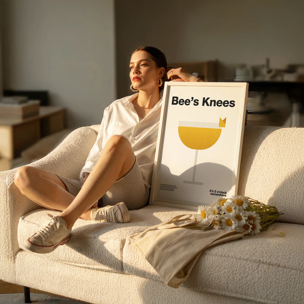 Woman sitting on a couch with a 'Bee's Knees' poster and flowers.