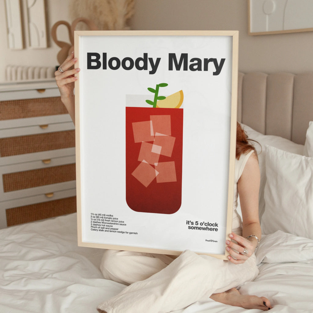 Person holding a framed print of a Bloody Mary cocktail in a bedroom setting.
