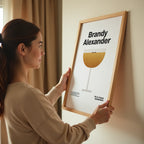 Woman holding a framed poster with 'Brandy Alexander' recipe on a wall.