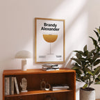 Framed poster titled 'Brandy Alexander' on a wooden shelf with books and decor.
