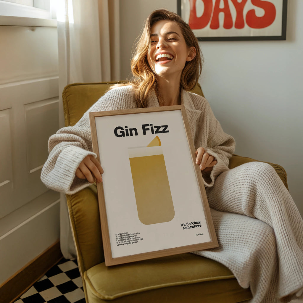 Woman holding a framed picture titled 'Gin Fizz' in a cozy room.