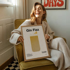 Woman holding a framed picture titled 'Gin Fizz' in a cozy room.