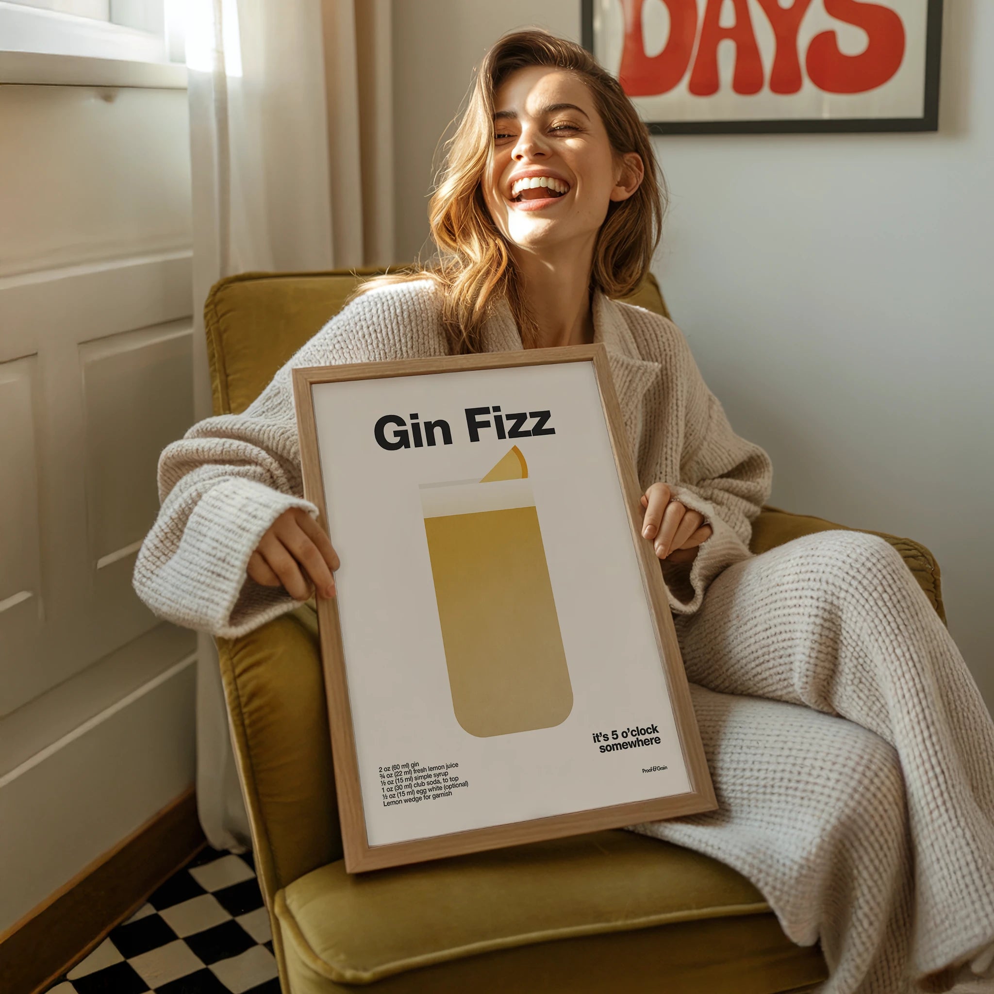 Woman holding a framed picture titled 'Gin Fizz' in a cozy room.
