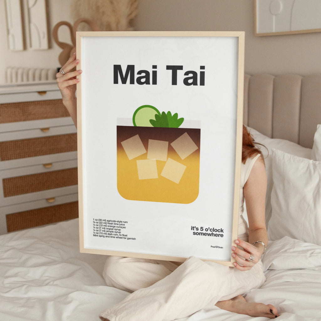 Person holding a framed poster with 'Mai Tai' design and text in a bedroom setting.