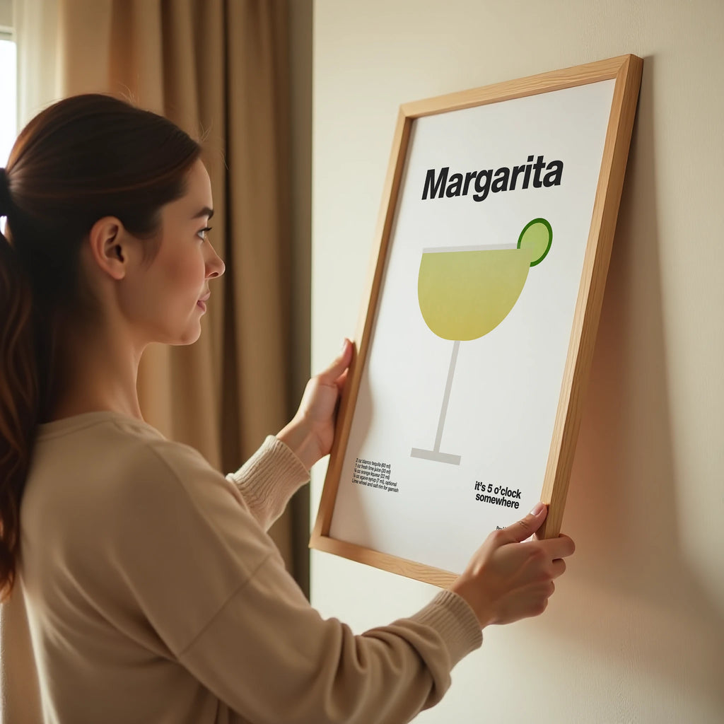 Woman holding a framed picture of a margarita illustration on a wall.
