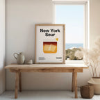 Framed poster of 'New York Sour' on a wooden table with a window in the background