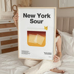 Person holding a framed poster titled 'New York Sour' in a bedroom setting.