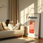 Woman reading a book on a beige sofa in a sunlit room with a 'Paloma' poster on the wall.