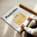 Person unrolling a poster with 'Penicillin' and a yellow capsule on a beige carpet.