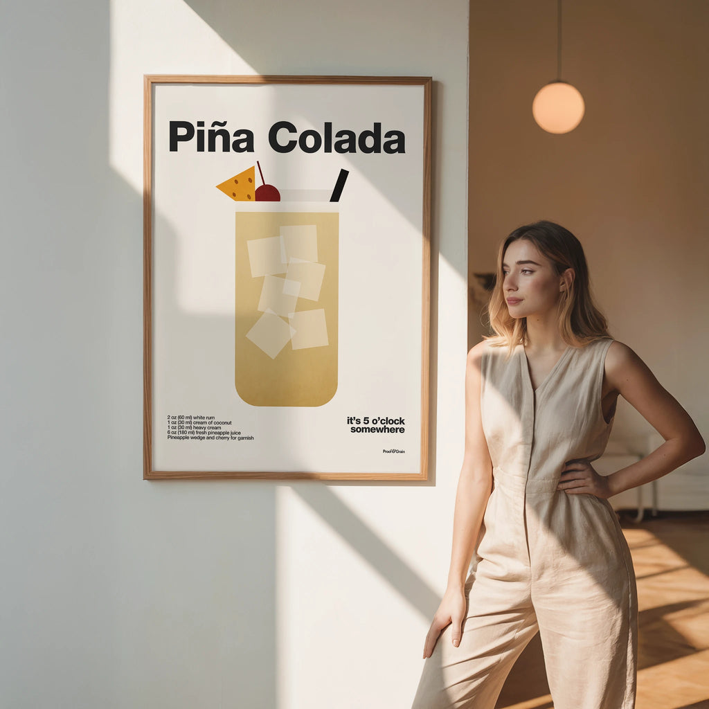 Woman standing next to a framed poster of a Pina Colada illustration in a room with warm lighting.
