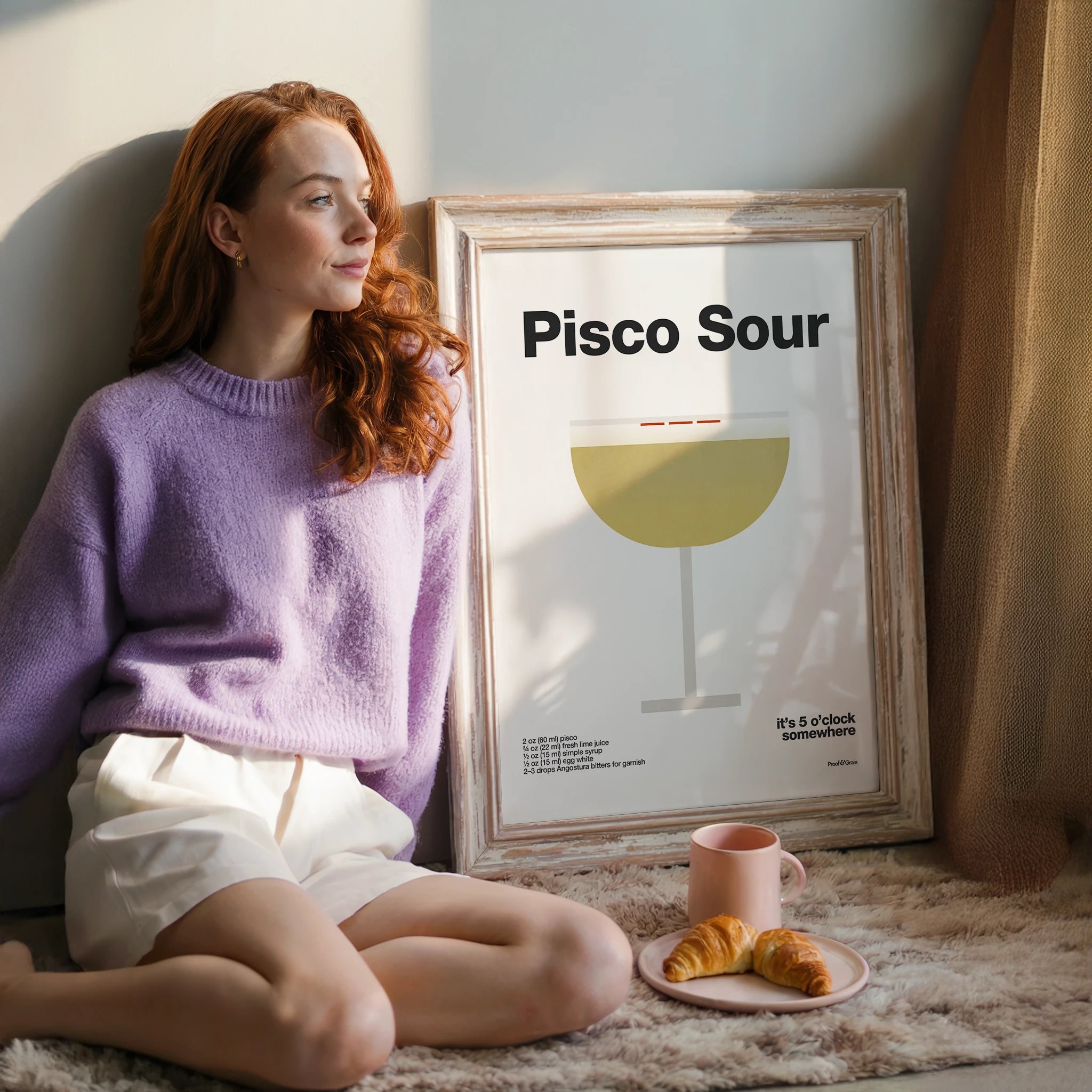 Woman sitting on a couch with a framed 'Pisco Sour' poster and a plate of food.