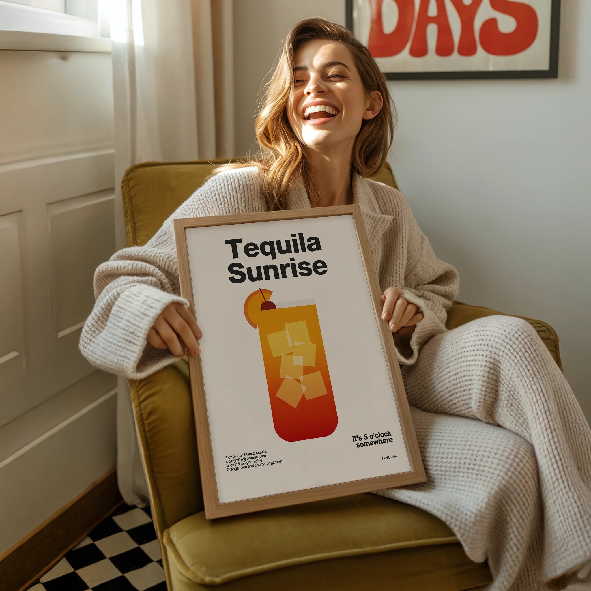 Woman holding a framed 'Tequila Sunrise' poster in a cozy room.