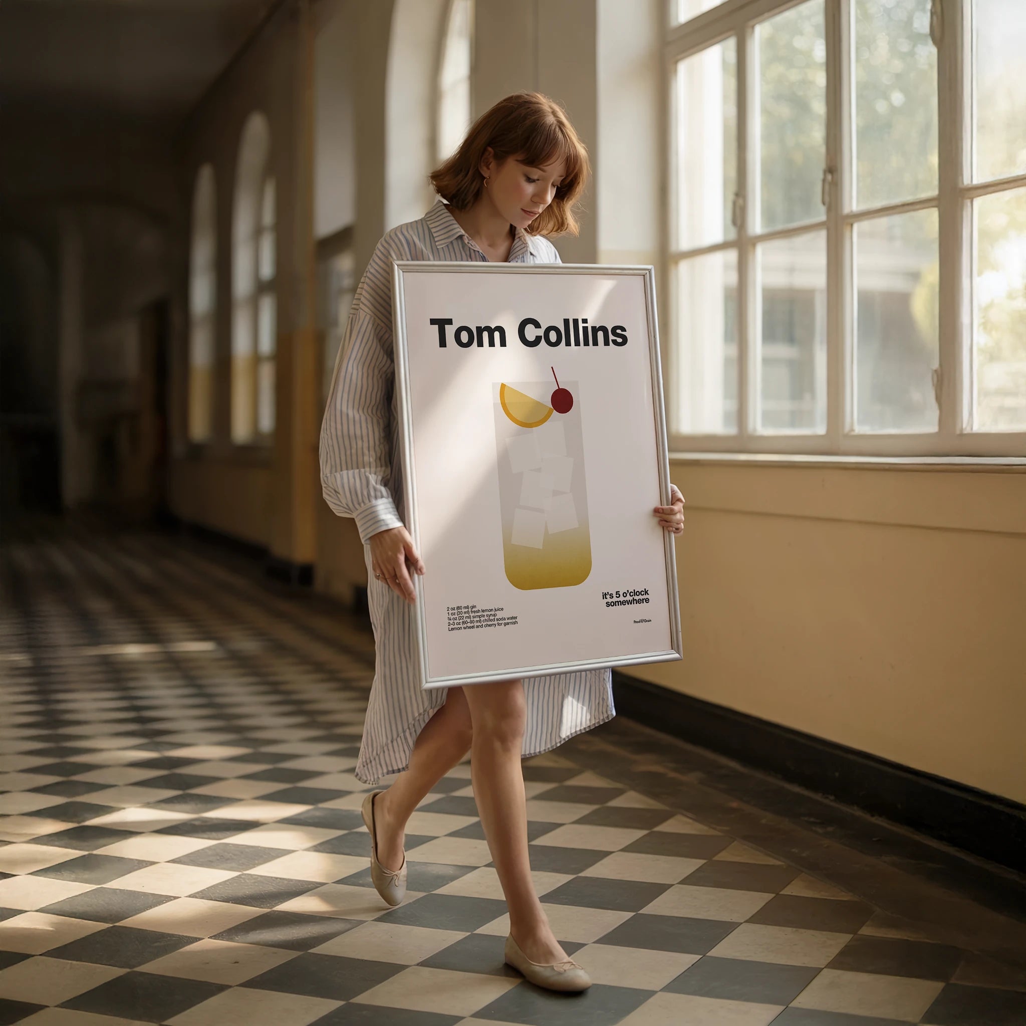 Woman holding a framed picture of a Tom Collins cocktail in a sunlit room.