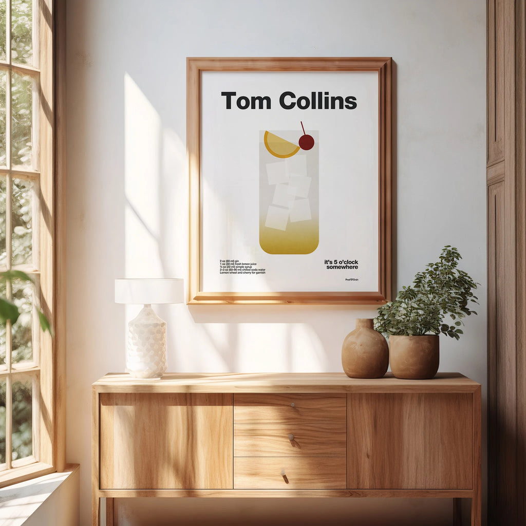 Framed 'Tom Collins' poster on a wall with a wooden cabinet and plants below.