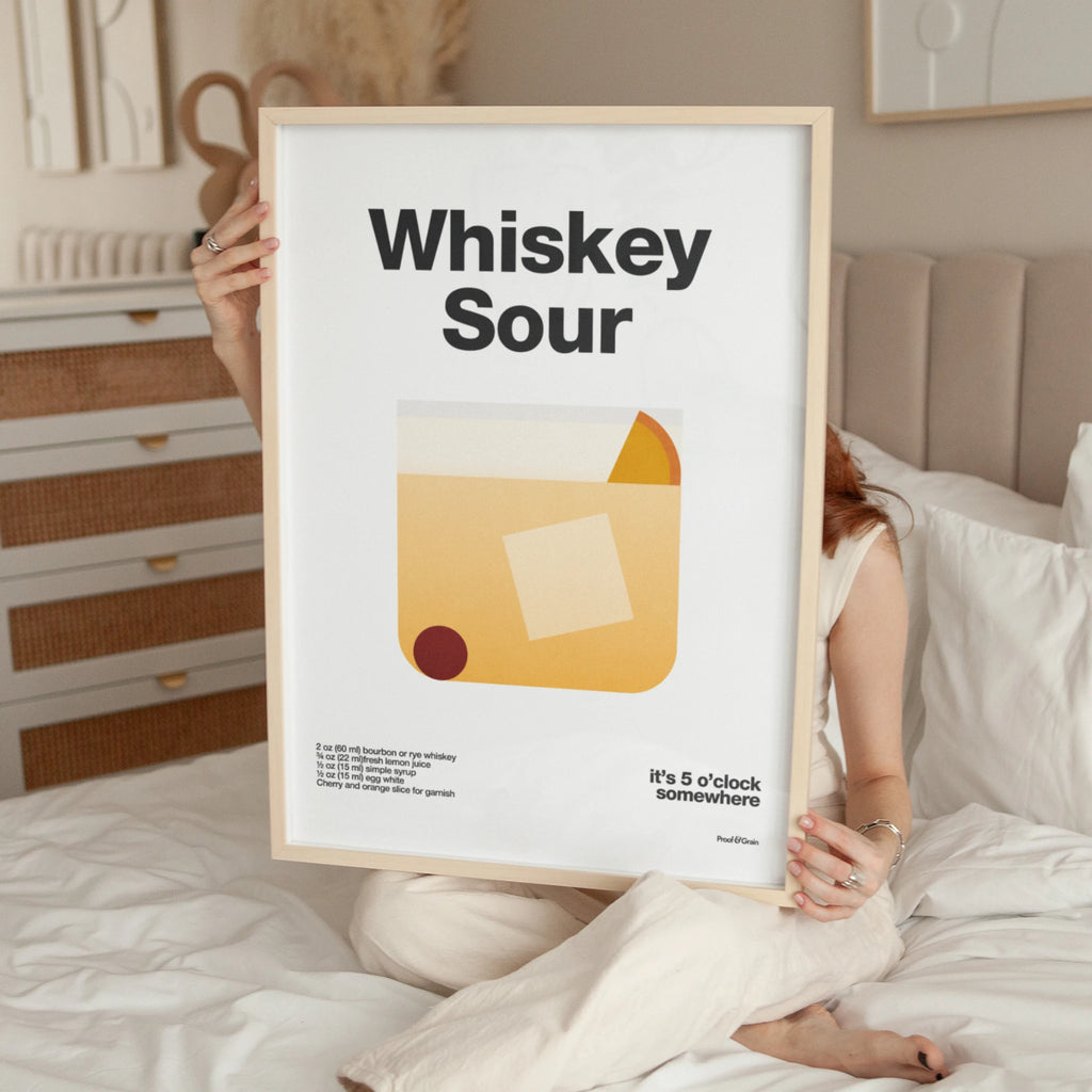 Person holding a framed poster with 'Whiskey Sour' design in a bedroom setting.
