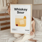 Person holding a framed poster with 'Whiskey Sour' design in a bedroom setting.